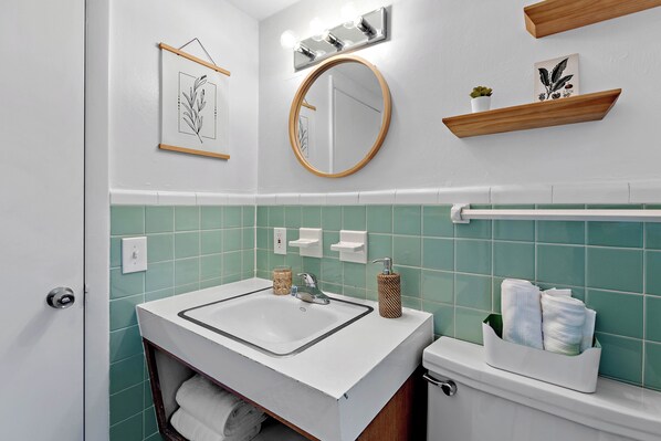 Un baño con un lavabo blanco, azulejos verdes, un espejo redondo y un estante de madera con una planta.