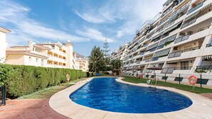 Una piscina rodeada por un solarium con baldosas, con un edificio que tiene balcones y un seto al fondo.