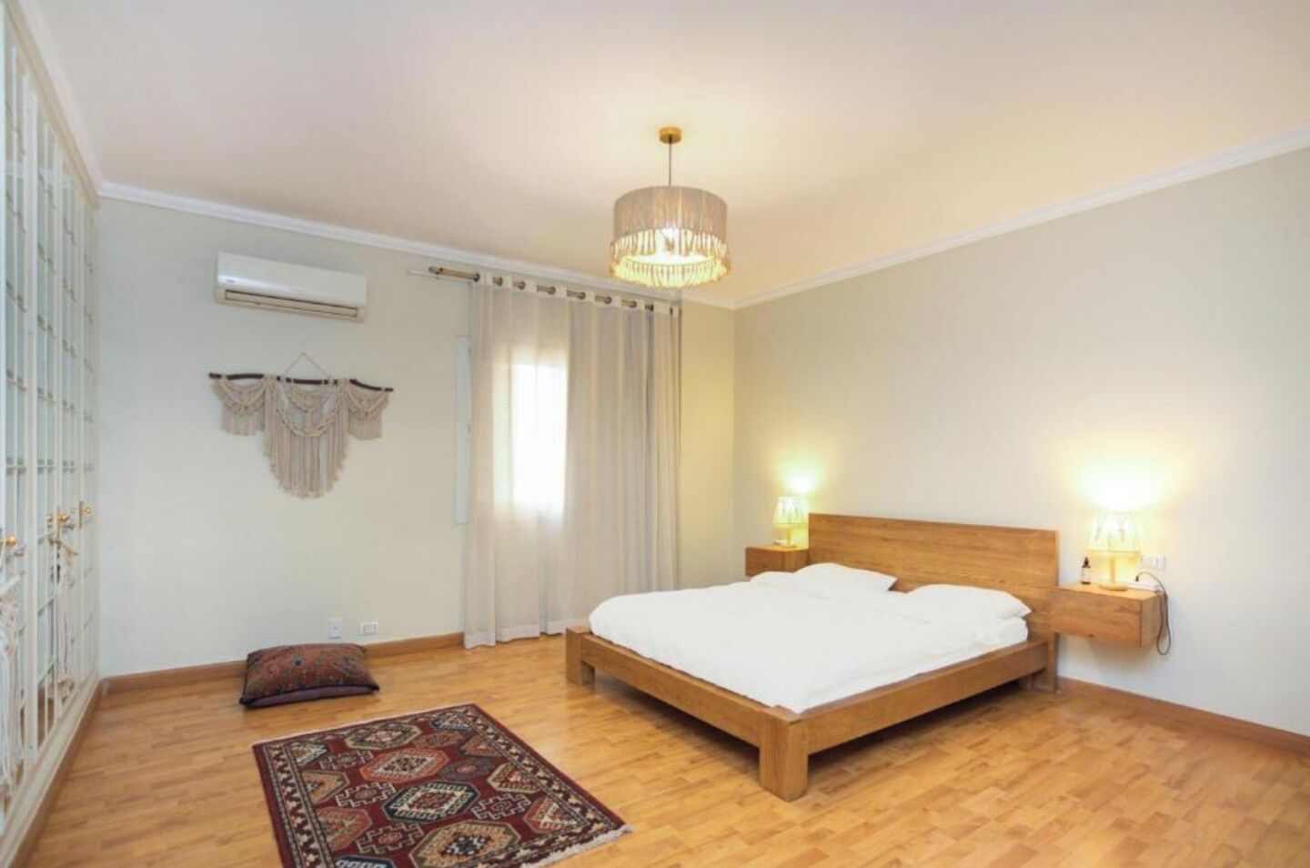 A bedroom with a wooden bed, two bedside lamps, a patterned rug, a macrame wall hanging, and an air conditioning unit.