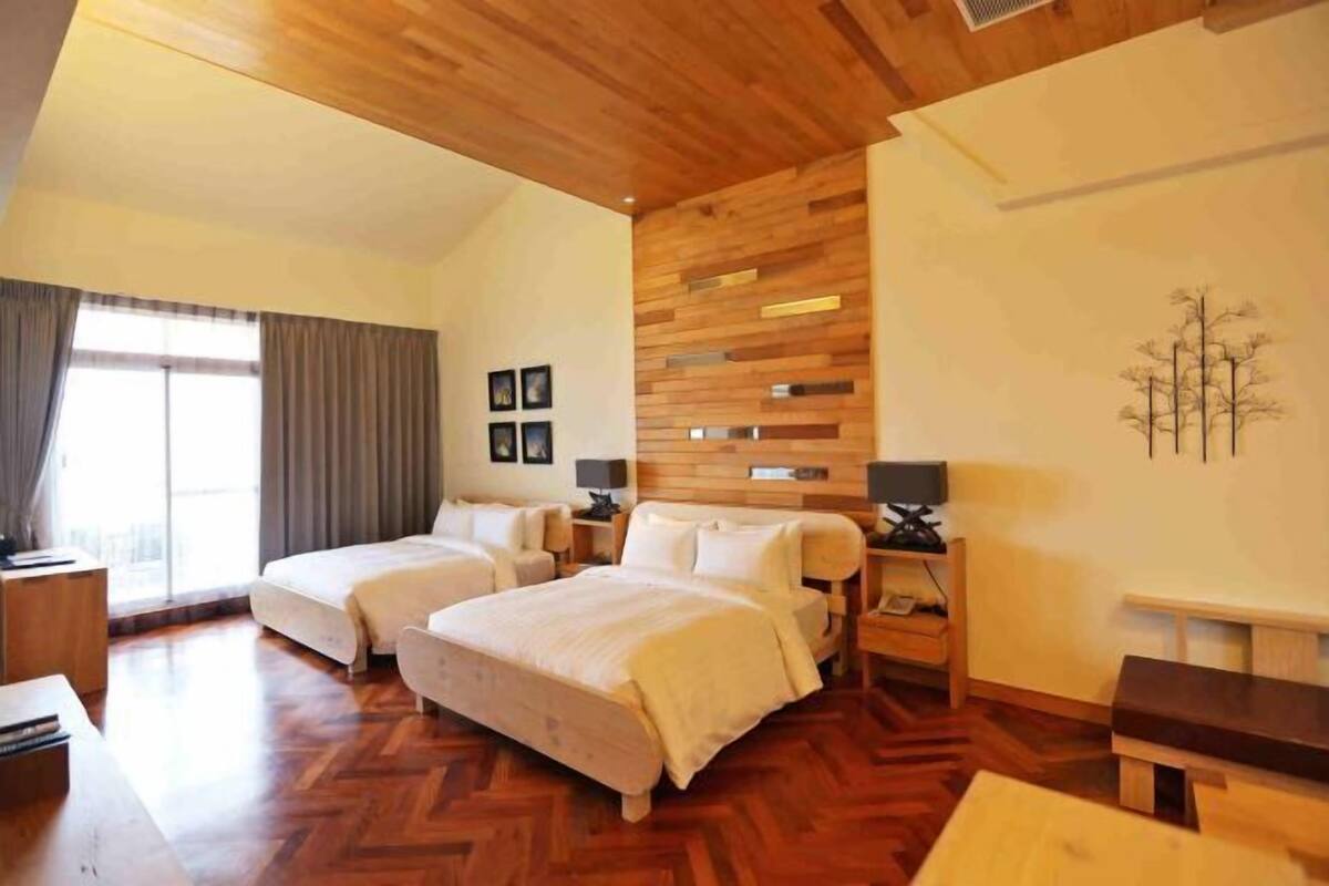 A hotel room with two beds, wooden flooring, a large window with curtains, and a wall-mounted TV.