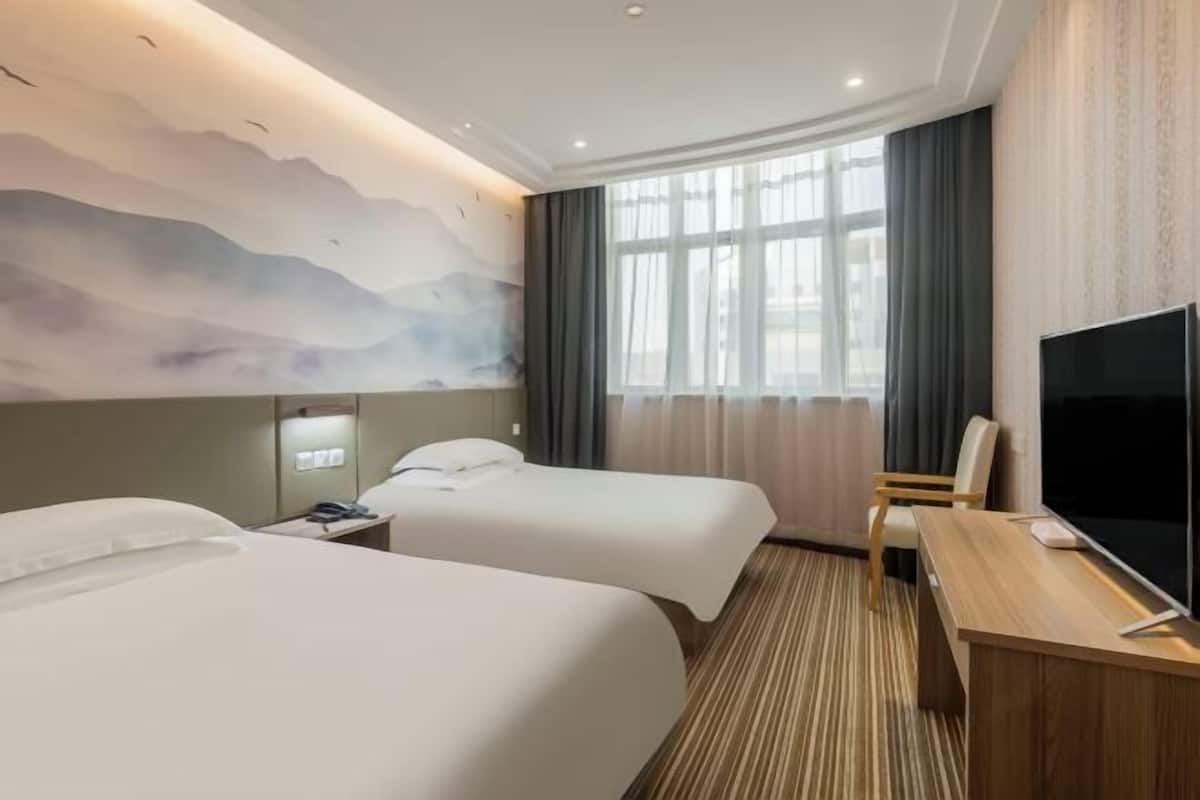 A hotel room with two beds, a desk, a TV, and a large window with curtains.