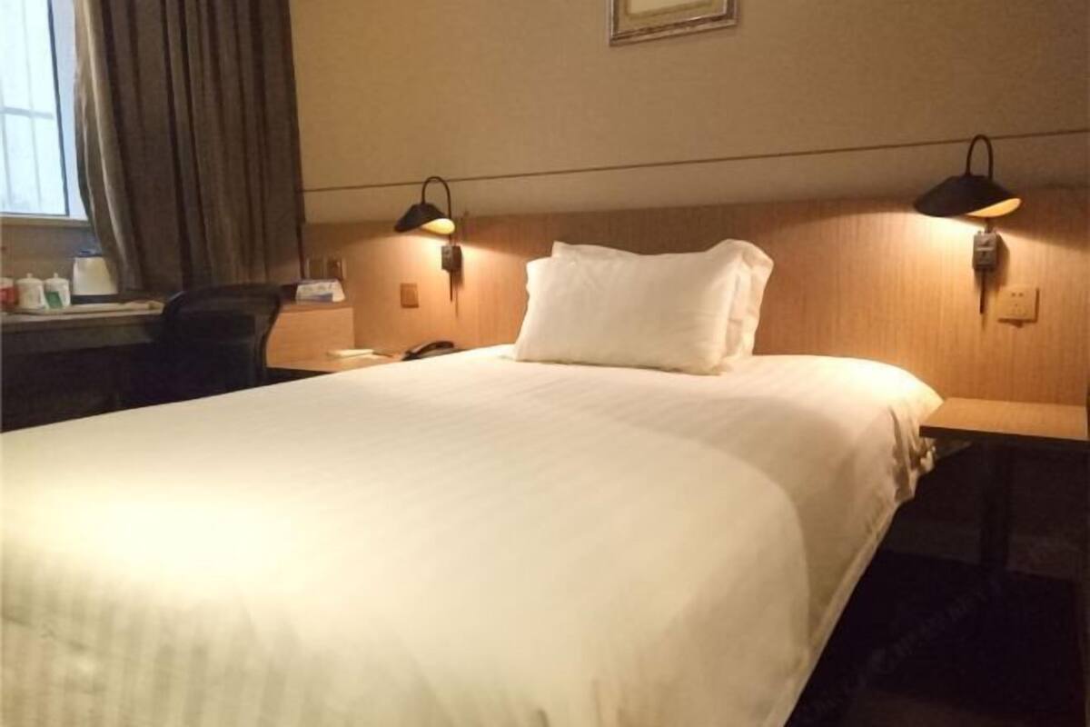 A hotel room with a bed, two bedside lamps, a desk with a chair, and a window with curtains.