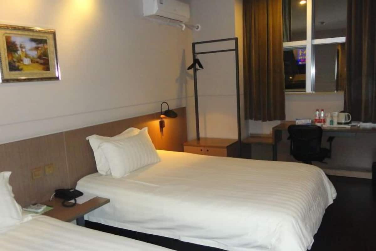 A hotel room with two beds, a desk, a chair, a window with curtains, a wall-mounted air conditioner, and a framed picture on the wall.