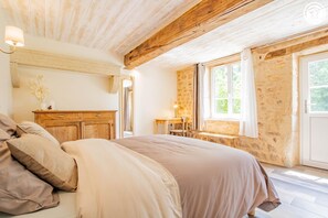 Ein Schlafzimmer mit Holzboden, einem Bett mit beigefarbener Bettwäsche, einem Nachttisch aus Holz, einem kleinen Tisch mit einer Lampe und einem Fenster mit Blick auf Grünflächen.