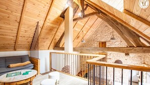 Un dormitorio tipo loft acogedor con techo inclinado, vigas de madera, pared de piedra, sofá gris, mesa redonda de madera y un taburete blanco acolchado.