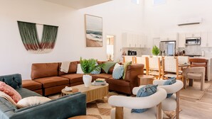 Un saló modern amb un sofà marró, un butàc armchair blau i una taula de centre. Hi ha una zona de cuina amb armaris blancs i una taula de menjador amb caires.
