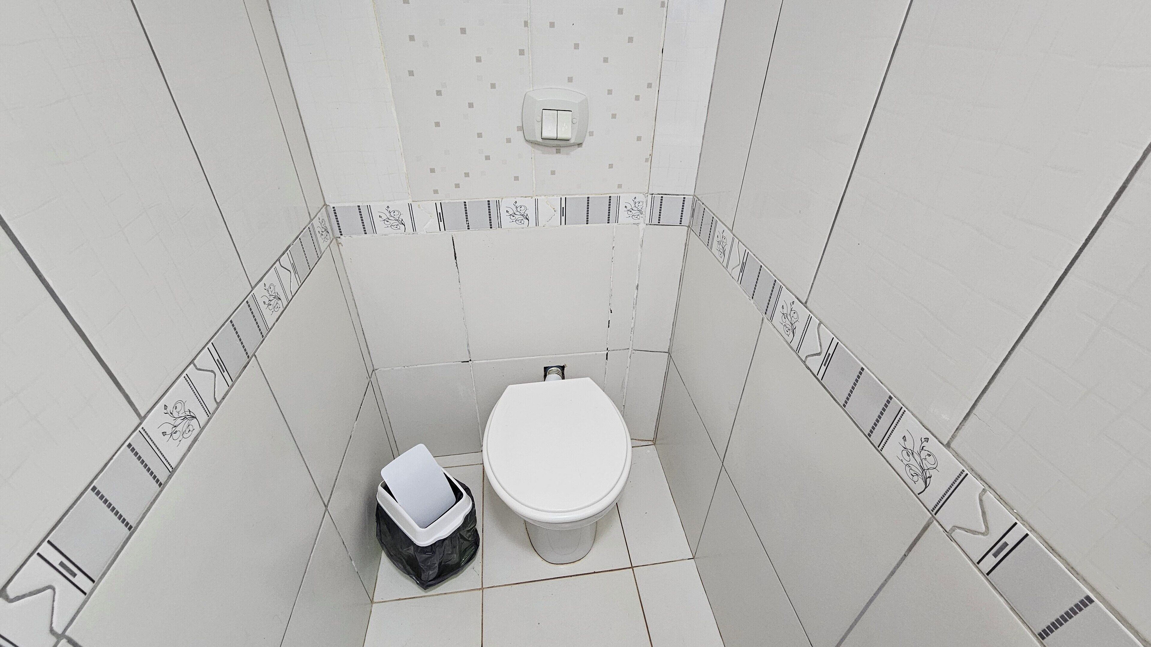 A small, tiled bathroom with a toilet, a trash bin, and a wall-mounted flush handle.