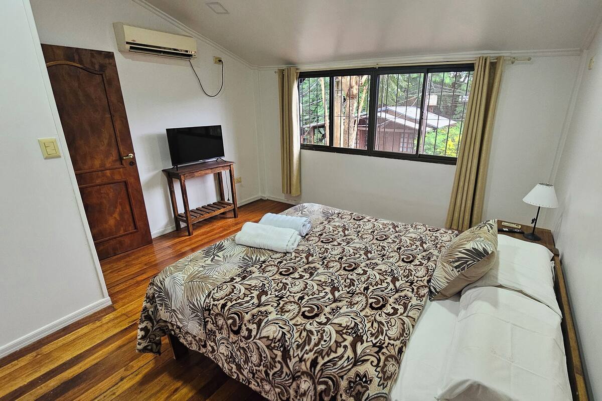 Ein Schlafzimmer mit einem Bett, einem Fernseher auf einem kleinen Tisch und einem Fenster mit Vorhängen.