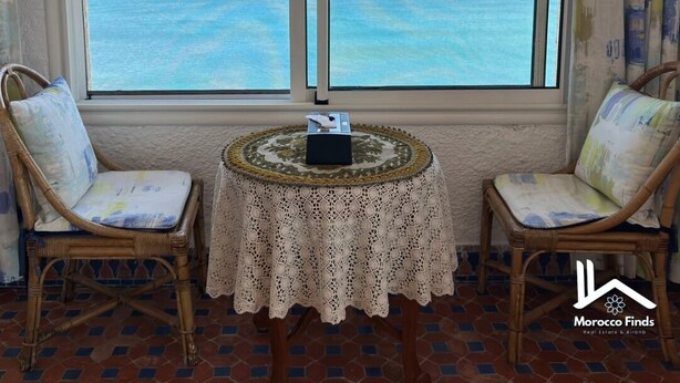 Une pièce dotée d’une grande fenêtre donnant sur la mer, de deux fauteuils en osier, d’une petite table ronde recouverte d’une nappe en dentelle et d’un sol carrelé.