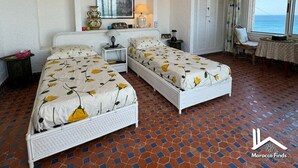 Une chambre avec deux lits, un ventilateur de plafond, un tableau, une fenêtre donnant sur un beau paysage et un sol carrelé.