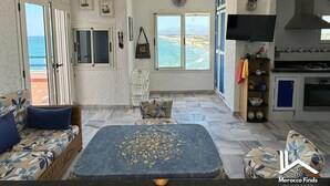 Una habitación con un ventanal grande con vista al mar, un ventilador de techo, una zona de cocina y una sala de estar con una mesa.