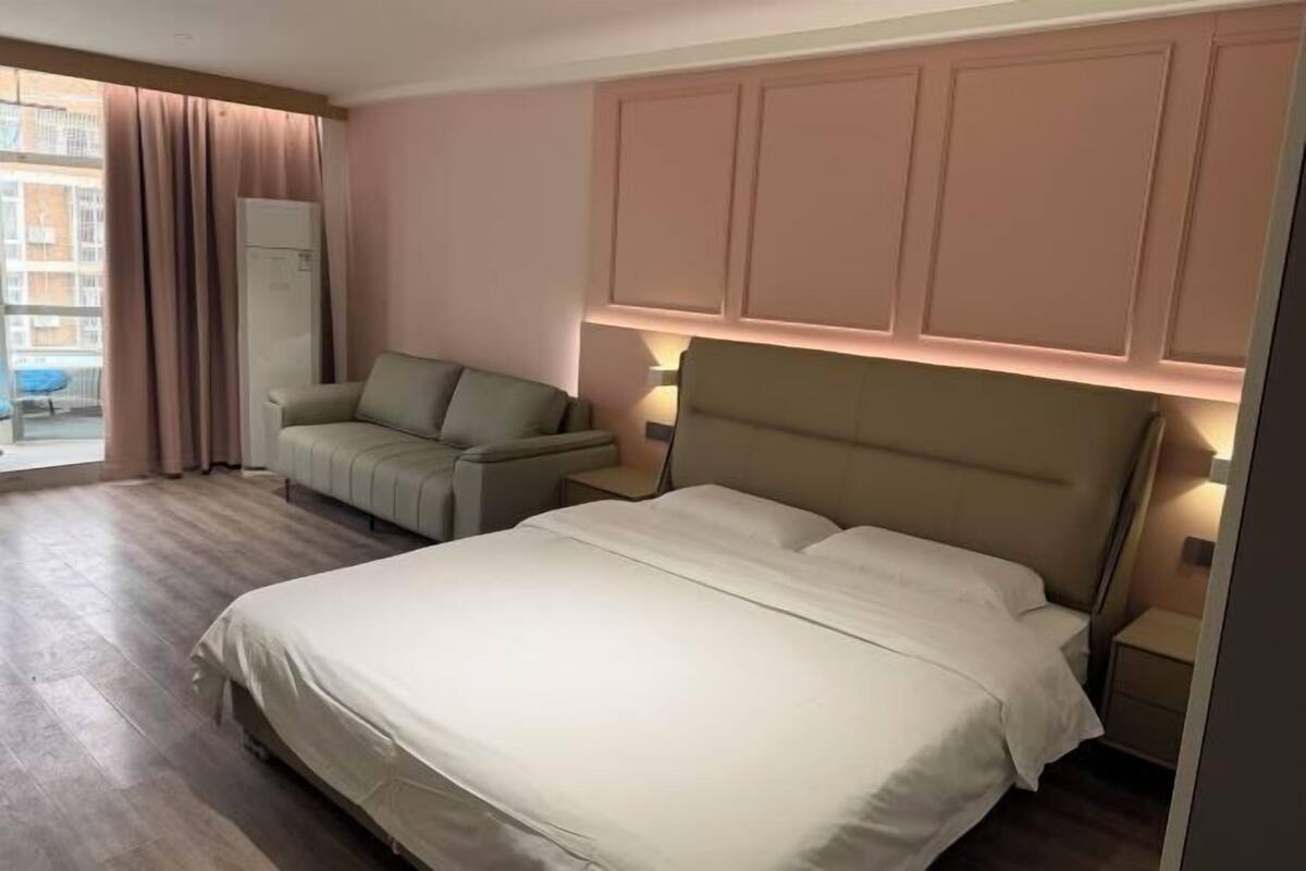 Ein Hotelzimmer mit Bett, Sofa, Nachttisch, Lampe, einem Fenster mit Vorhängen und Blick auf Gebäude.