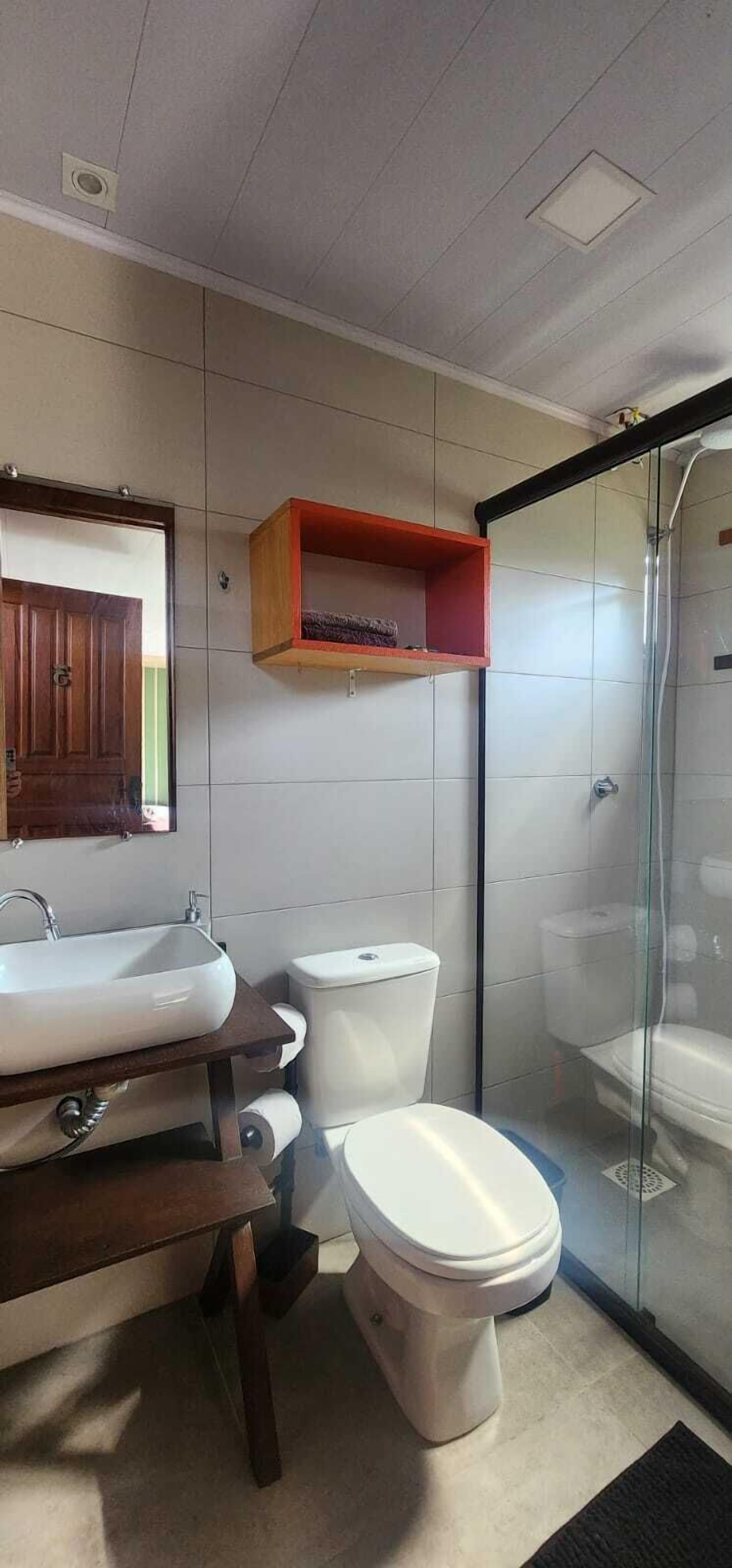 A modern bathroom with a wooden vanity, a glass shower enclosure, and a small shelf above the toilet.