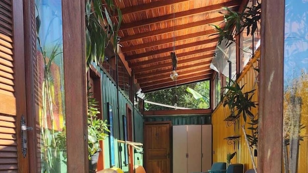 A modern interior with a wooden ceiling, a central seating area with colorful chairs, and a view of the outdoors. - Moçamba LODGE Contêiner (Florianópolis)