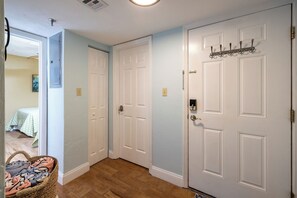 A hallway with two white doors, a wicker basket with clothes, and a bed with a green blanket in the background. - Breathtaking Direct Oceanfront Views! Huge Community Pool, Private Beach Access! (New Smyrna Beach)