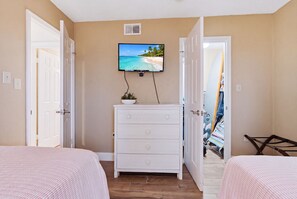 Une chambre avec deux lits, une commode, un téléviseur fixé au mur et une porte donnant sur une autre pièce.