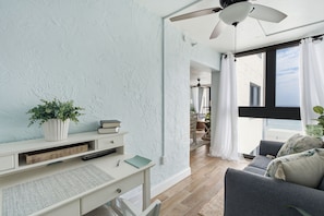 Ett modernt vardagsrum med ett skrivbord, en takfläkt, ett stort fönster med gardiner och utsikt över omgivningarna.