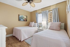 Een slaapkamer met twee bedden, een plafondventilator, een raam met gordijnen, een nachtkastje en een aan de muur hangend schilderij.