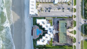 An aerial view of a coastal hotel with a swimming pool, tennis courts, and a parking area. - Breathtaking Direct Oceanfront Views! Huge Community Pool, Private Beach Access! (New Smyrna Beach)