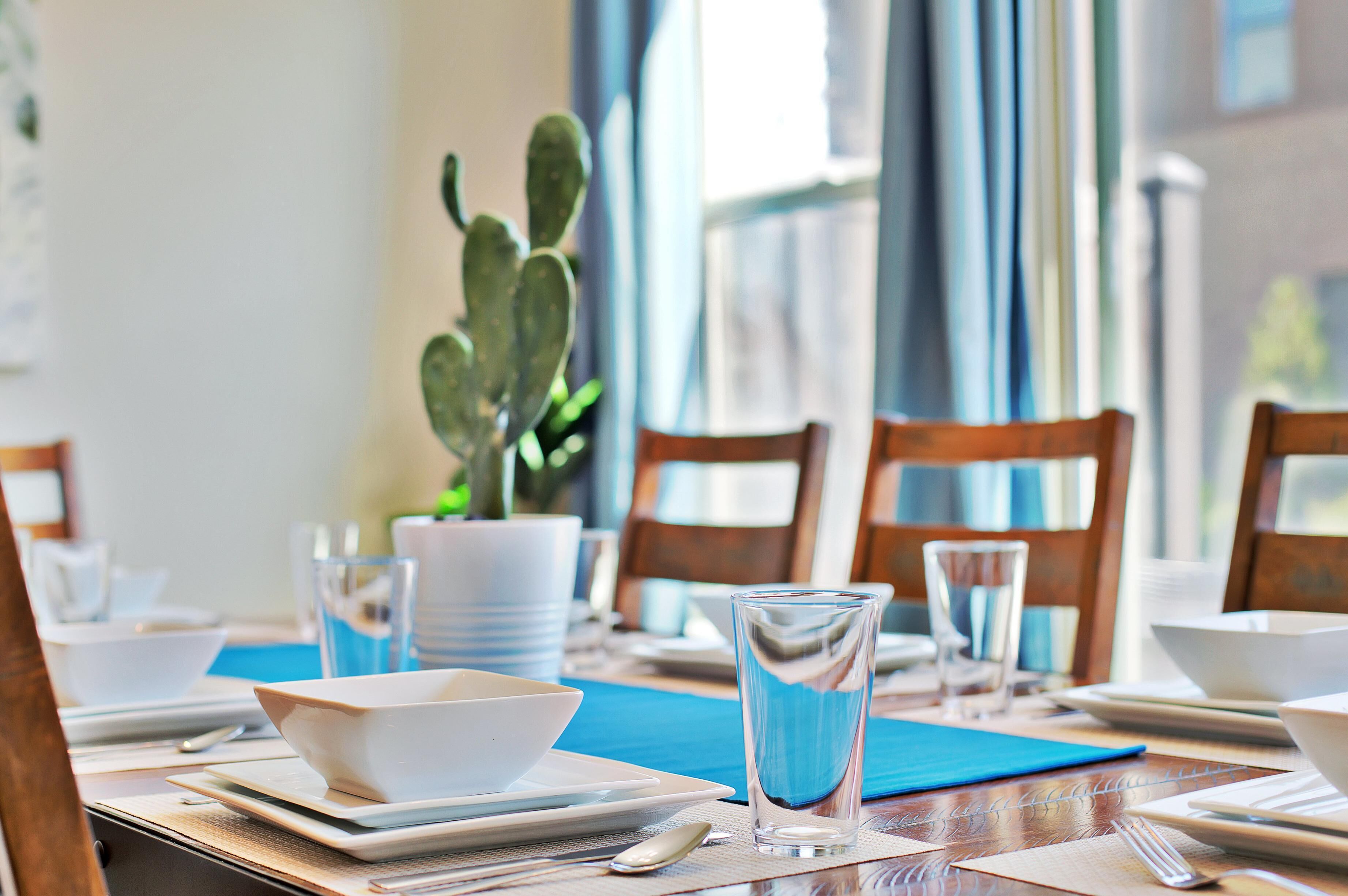 A dining table set with white plates, bowls, and clear glasses, with a cactus plant in the background.