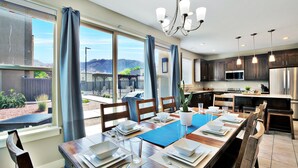 A modern kitchen with stainless steel appliances, a dining area with a blue table runner, and large windows offering a view of the outdoors. - Entrada at Moab 619 (Moab)