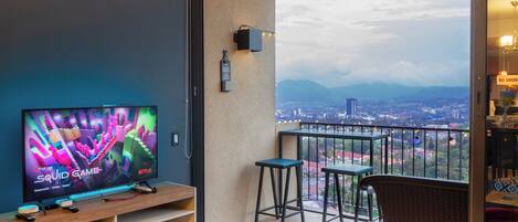 Un balcón con televisor, taburetes de bar y vista a un paisaje urbano.