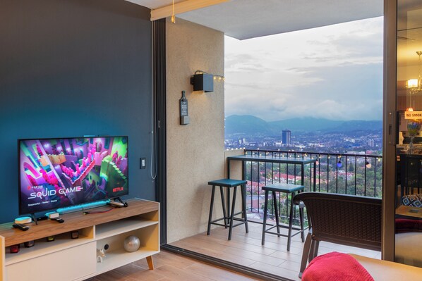 A balcony with a TV, bar stools, and a view of a cityscape.