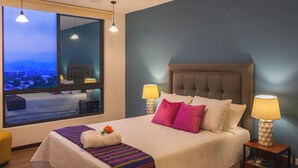 A neatly made bed with a white comforter and colorful pillows, a bedside table with a lamp, and a large window with a city view. - Deluxe, City views, Rooftop pool, upscale location (San Salvador)