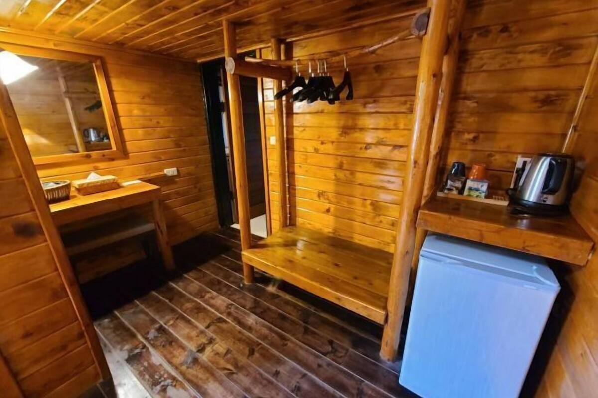 A wooden cabin interior with a wooden bench, a small table, a mirror, and a refrigerator.