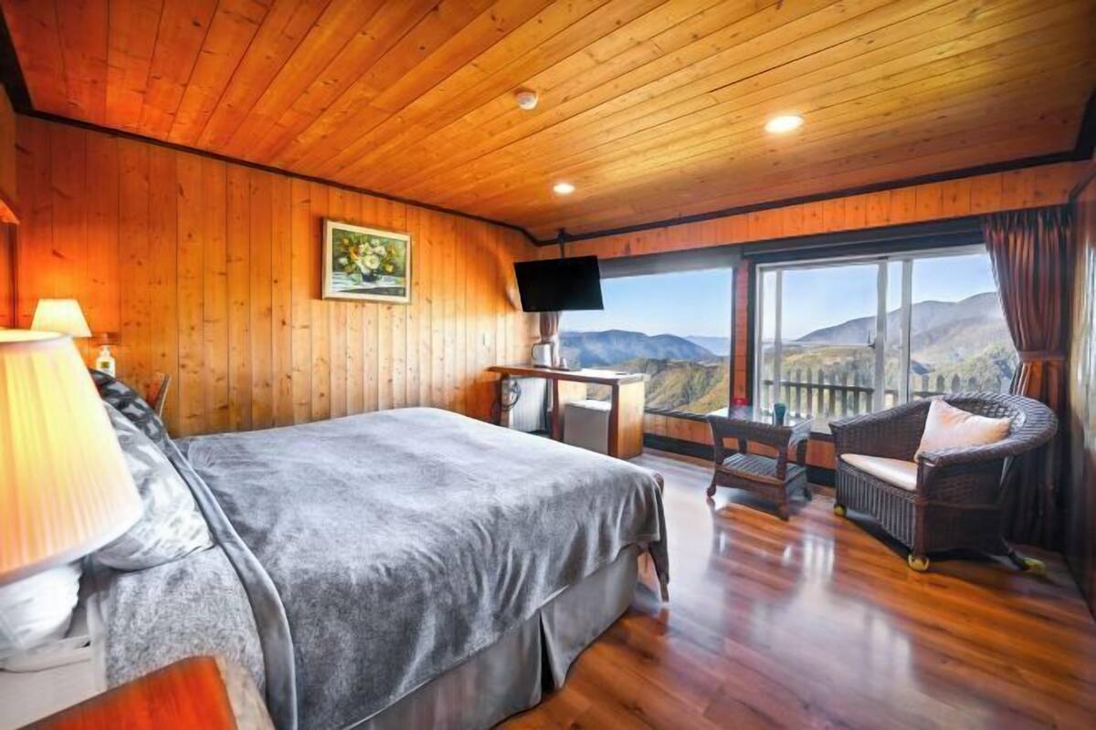 A wooden-paneled room with a bed, a desk, a chair, and a large window offering a mountain view.
