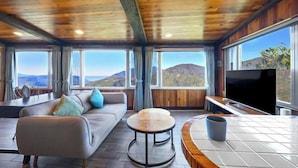 A modern living room with a wooden ceiling, large windows offering a mountain view, a sofa with cushions, a round coffee table, and a flat-screen TV. - Chingjing Top Cloud Resort (Nantou)