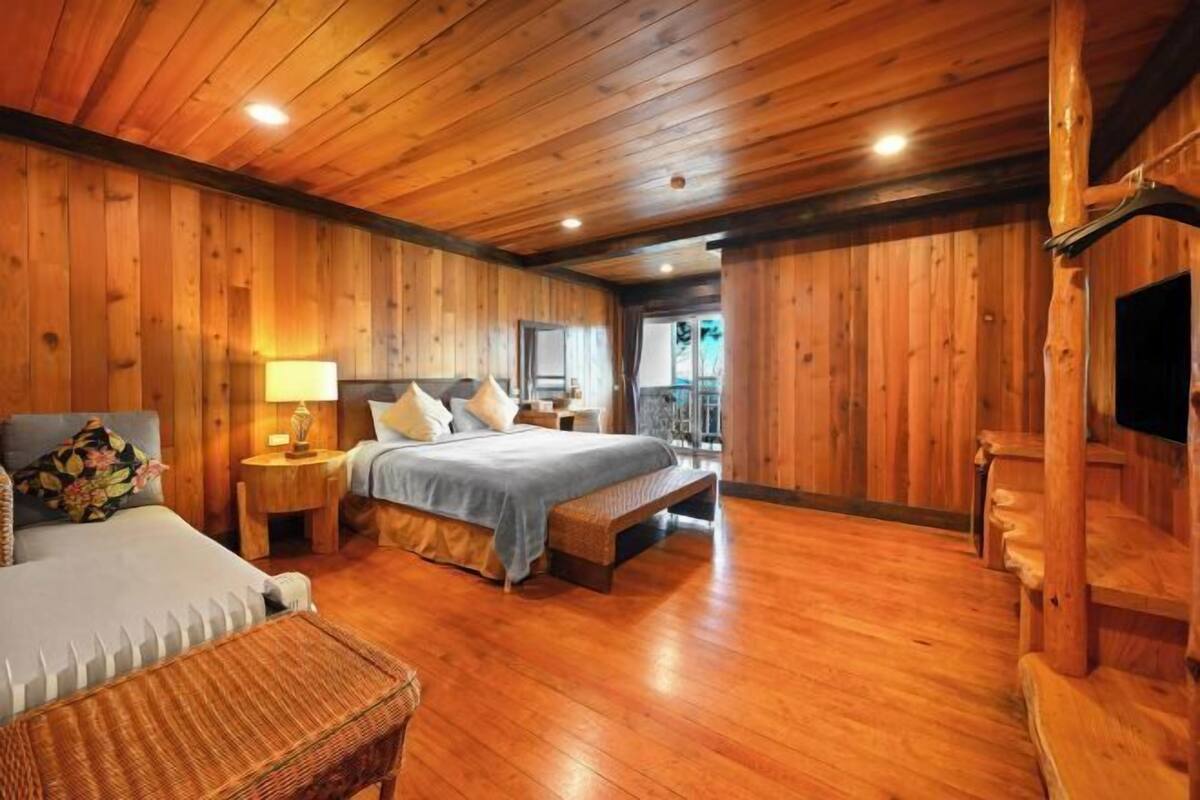 A wooden cabin bedroom with a bed, bedside table, lamp, and a view of the outside.