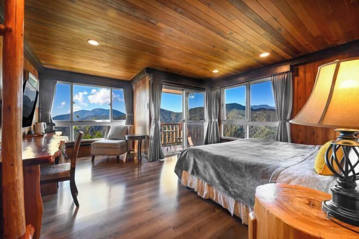 A bedroom with a large bed, wooden ceiling, and multiple windows offering mountain views.