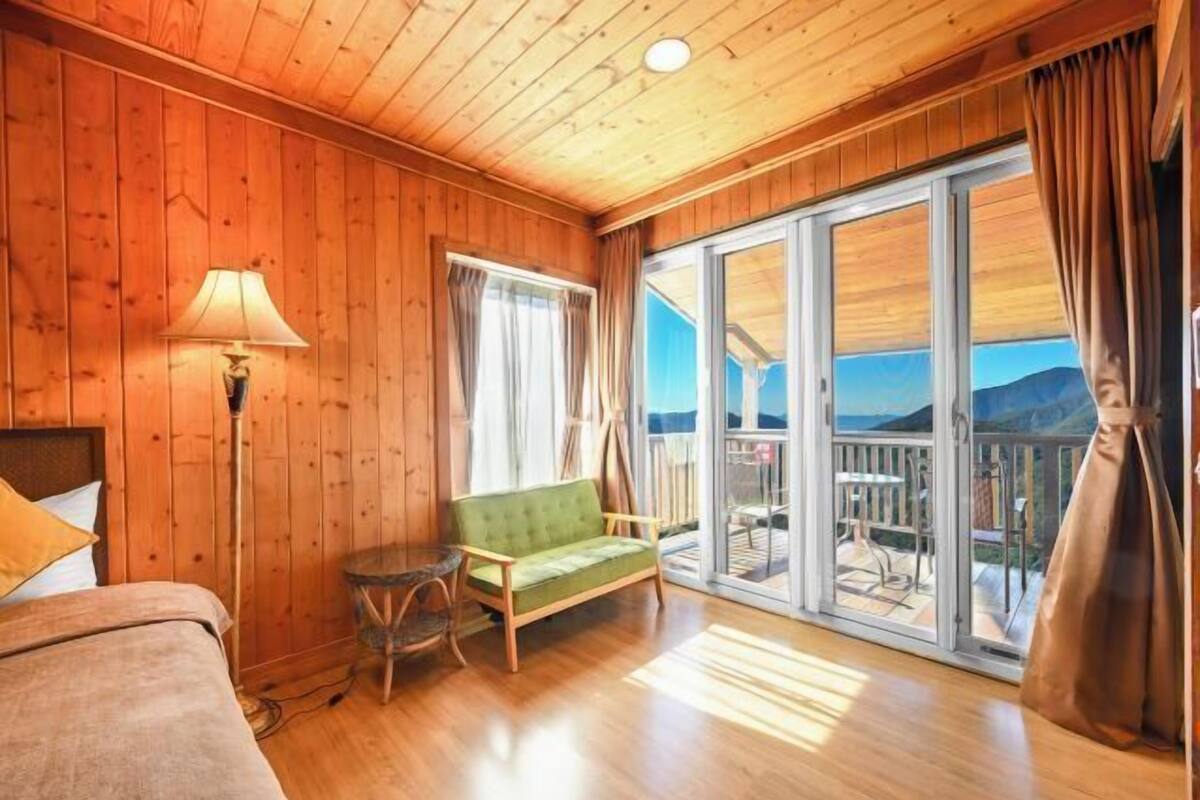 A wooden cabin room with a bed, a green sofa, a small table, and a balcony with a view of the mountains.