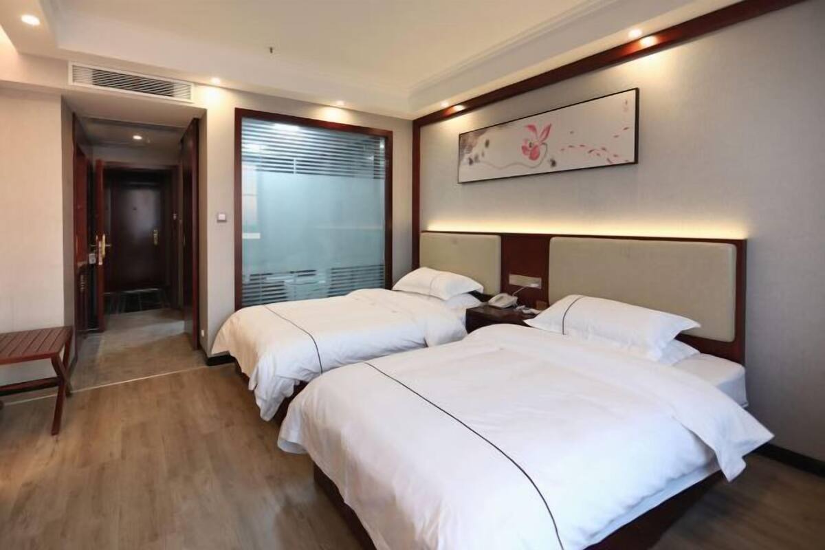 A hotel room with two beds, a bedside table, a wall-mounted artwork, and a door leading to a hallway.