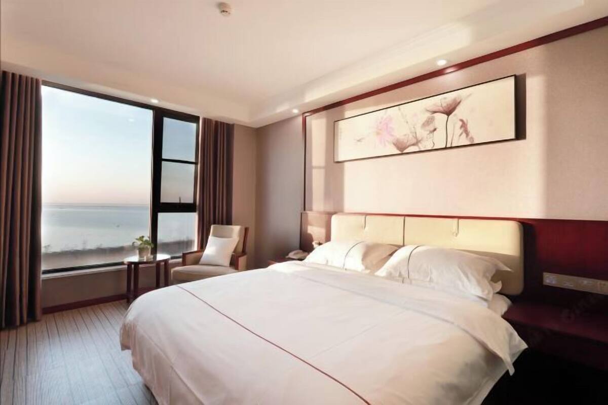 Habitación de hotel con una cama grande, una silla, una mesita de noche, un cuadro en la pared y vista al mar a través de un ventanal amplio.