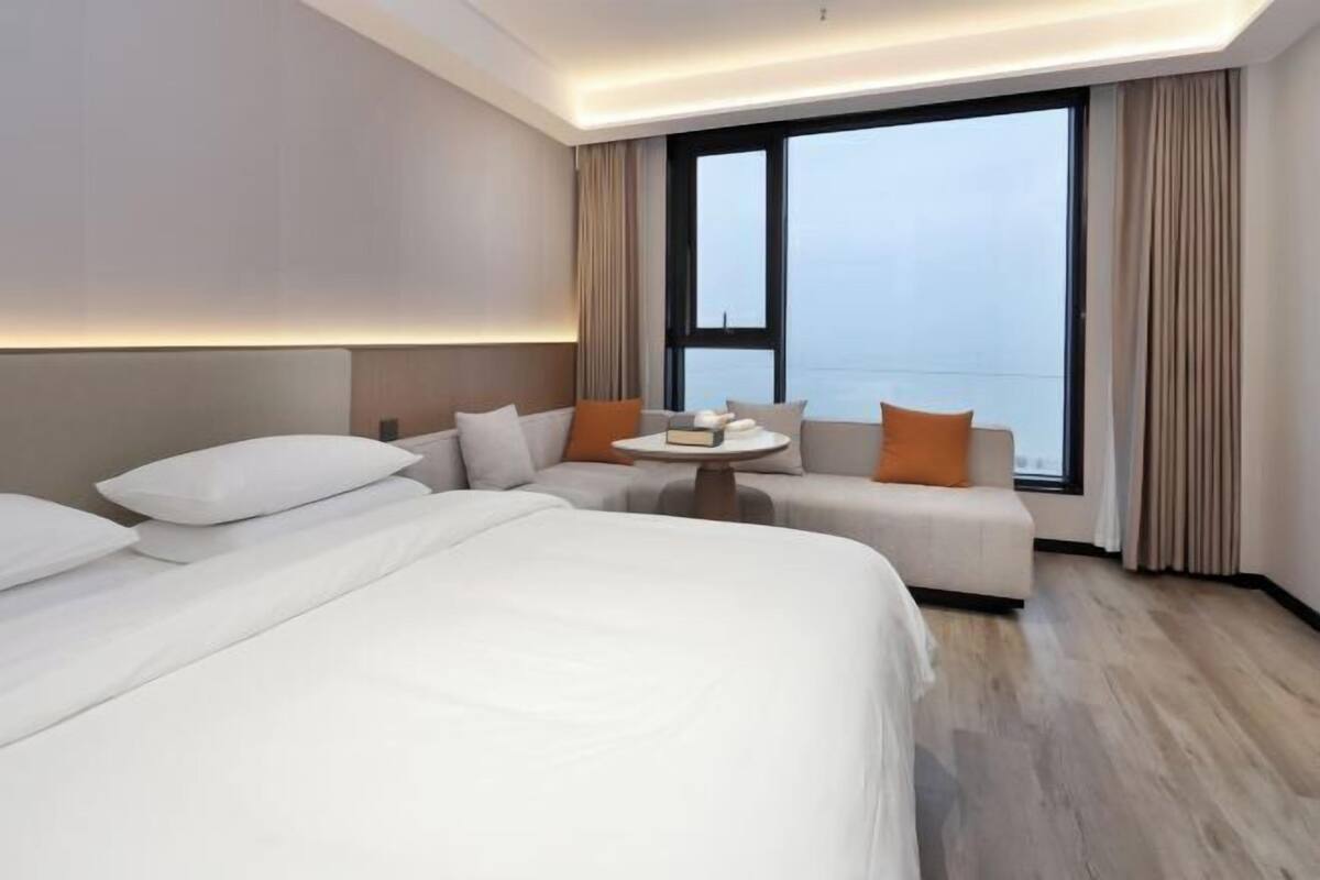 Habitación de hotel moderna con una cama grande, un sofá y una mesita. Cuenta con un ventanal con cortinas y una paleta de colores neutros.