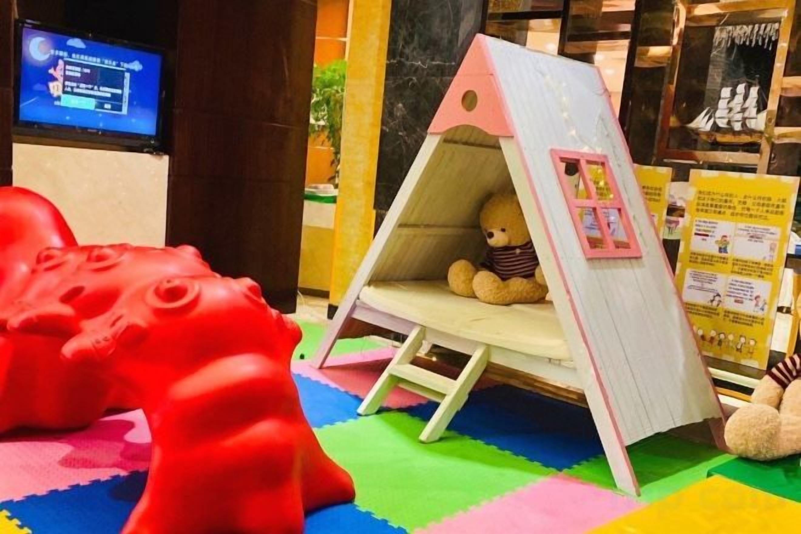 A play area with a red toy dinosaur, a small tent with a teddy bear, and a television screen in the background.