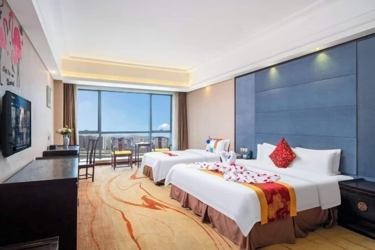 Ein Hotelzimmer mit zwei Betten, einem Schreibtisch, einem Stuhl, einem Fernseher und einem großen Fenster mit Blick auf die Stadt.