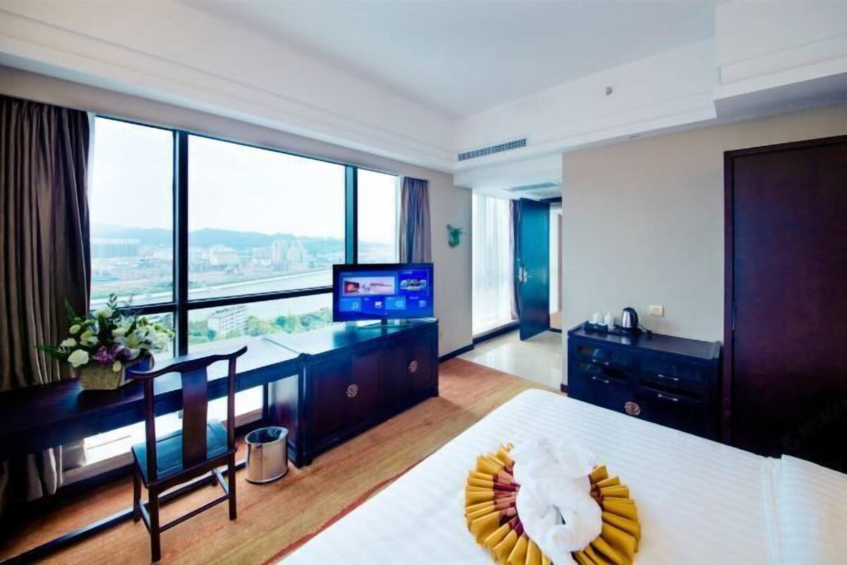 Ein Hotelzimmer mit großem Fenster, einem Bett mit Zierkissen, einem Schreibtisch mit Fernseher und einer Minibar.