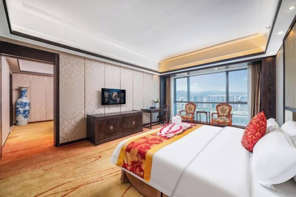 Ein Hotelzimmer mit einem großen Bett, einem Fernseher, einem Schreibtisch und Blick auf die Stadt.