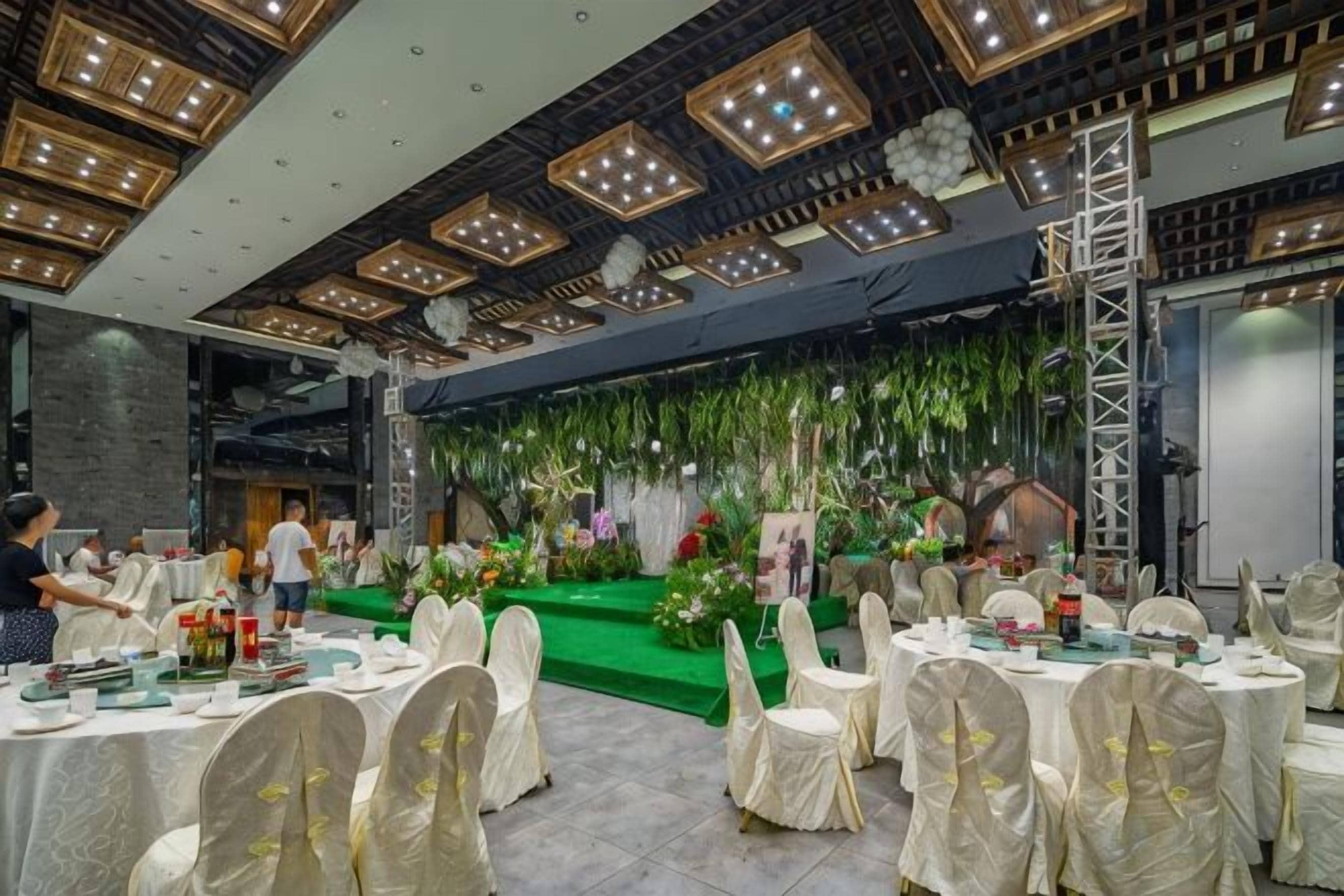 A banquet hall with round tables covered in white tablecloths and gold sashes, a stage with floral decorations, and a modern ceiling with recessed lighting.