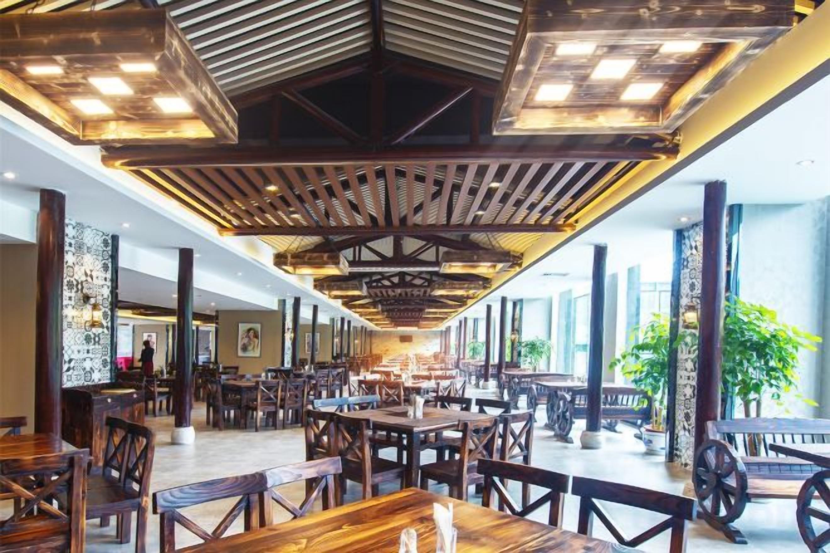 An indoor dining area with wooden tables and chairs, a high ceiling with exposed beams, and large windows allowing natural light.