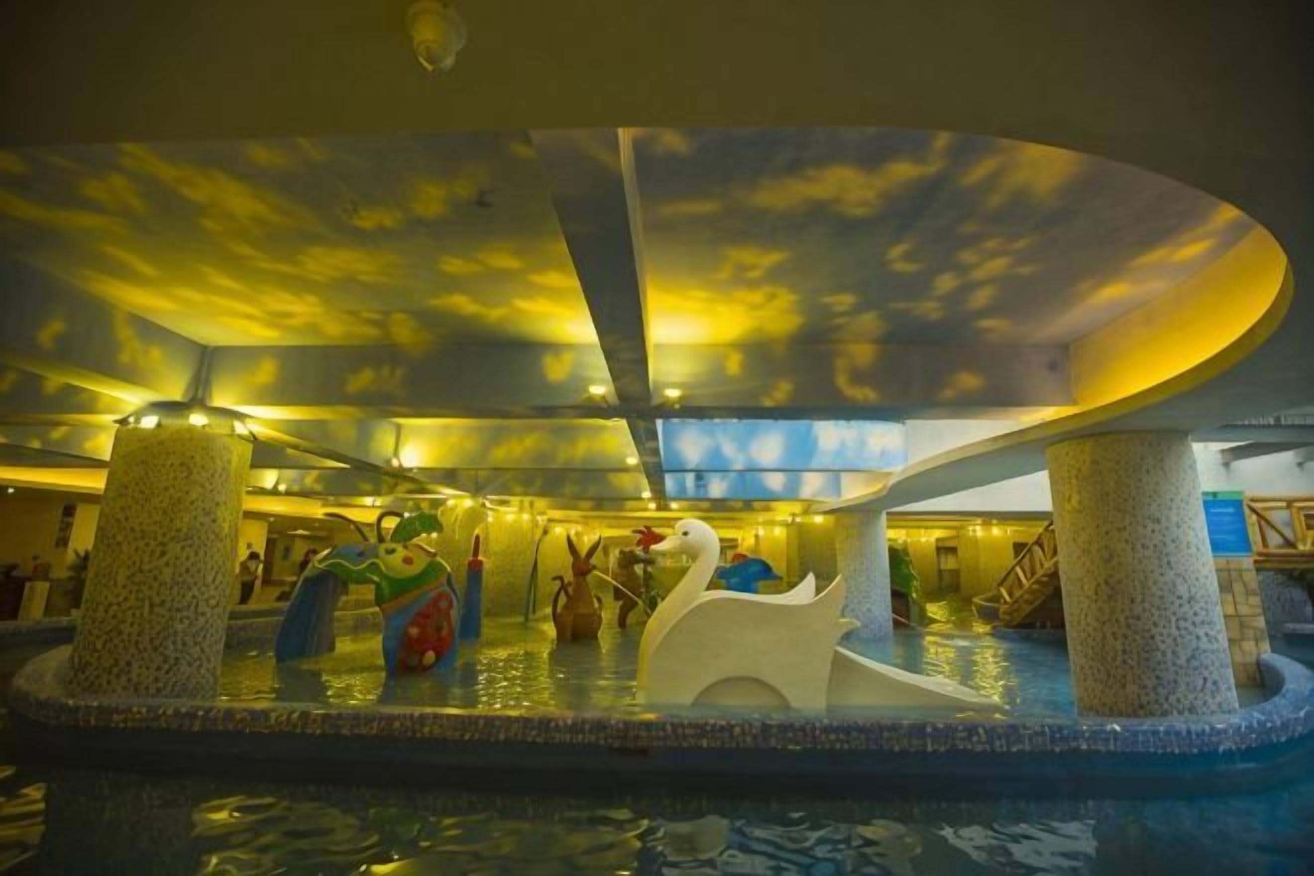 An indoor water feature with a swan sculpture, a colorful fish sculpture, and a ceiling with a cloud pattern.