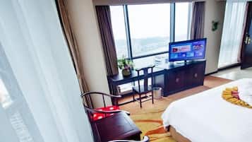 Habitación de hotel con una cama grande, un escritorio con televisor, una silla y vistas a un paisaje urbano a través de amplias ventanas.