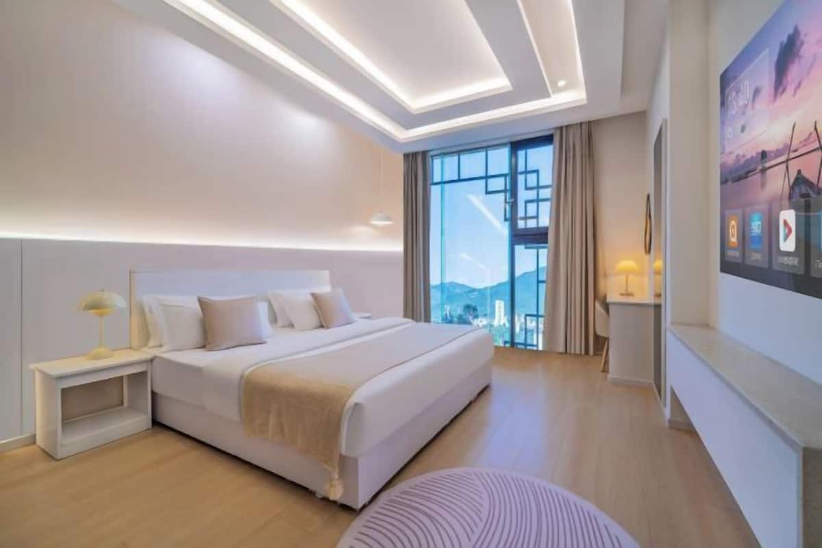 A modern hotel room with a large bed, a nightstand, a wall-mounted TV, and a large window offering a city view.