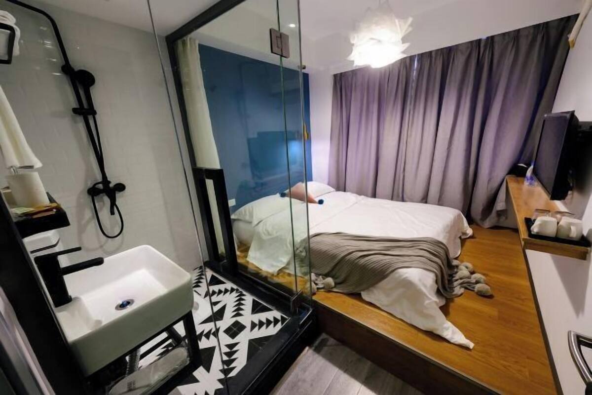A modern hotel room with a large mirror, a bed with a white comforter, a wooden floor, a built-in sink, and a television on a wooden shelf.