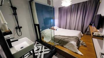 A modern hotel room with a large mirror, a bed with a white comforter, a wooden floor, a built-in sink, and a television on a wooden shelf.