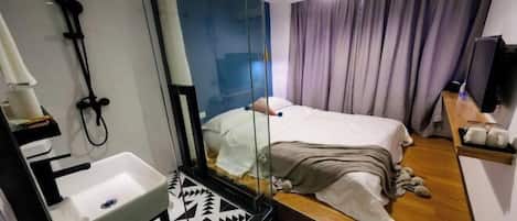Une chambre d’hôtel moderne avec un grand miroir, un lit recouvert d’une couette blanche, un plancher en bois, un lavabo intégré et un téléviseur posé sur une étagère en bois.