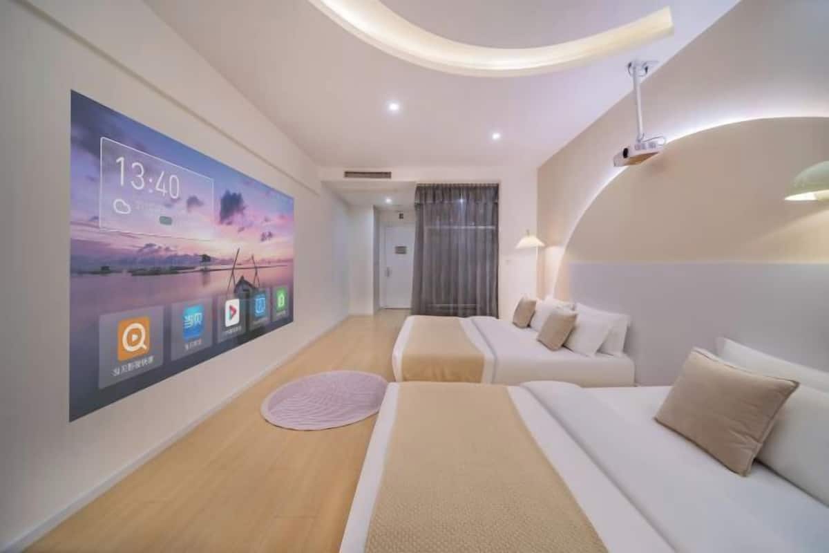 A modern hotel room with a large digital clock displaying the time and weather, a bed with beige bedding, a sofa with white cushions, and a round rug on the wooden floor.